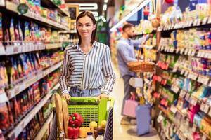 Everyday Shopping. Portrait Of Casual Female Customer Walking With Trolley Cart Between The Aisles In Local Store. Young Woman Buying Healthy Food In Shop, Looking At Shelves, Selective Focus photo