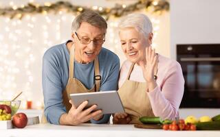 Joyful Senior Couple Using Tablet Making Call Standing In Kitchen On Christmas Eve. Remote Xmas Celebration Online. Older Spouses Chatting With Distant Family Using Computer photo