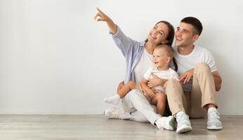 Happy Young Family Of Three With Infant Baby Sitting On Floor Near White Wall And Pointing Aside At Copy Space, Parents And Child Demonstrating Free Place For Your Design Or Advertisement, Panorama photo