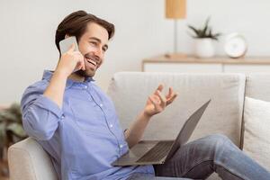 Cheerful Guy Talking On Phone Sitting With Laptop Computer On Sofa At Home. Modern Communication And Lifestyle. Happy Man Chatting On Cellphone And Laughing Indoors photo