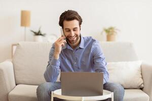 Business Talk. Man Talking On Cellphone Using Laptop Working Remotely From Home, Sitting On Sofa Indoor. Freelancer Speaking By Phone While Works On Computer. Freelance And Distance Work Concept photo