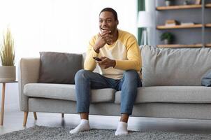 Watching TV at home. Happy african american man having rest, switching channels with remote controller and smiling, empty space. Black guy enjoying his weekend, watching TV in living room photo