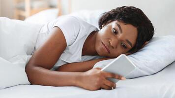 Frustrated young black woman looking at smartphone screen while laying in bed before sleeping, feeling lonely or depressed, closeup. Upset african american lady waiting for call from lover photo