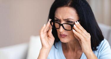 Poor Eyesight Concept. Closeup Portrait Of Mature Woman Squinting, Trying To Look Closer Through Eyeglasses At Something, Having Near Vision Problem At Home, Touching Eyewear At Home photo