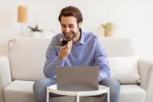 Smiling Businessman Talking By Phone In Loudspeaker Mode Or Using Voice Assistant On Smartphone Sitting At Laptop Computer On Couch Indoors. Gadgets And Modern Lifestyle, Voice Search Application photo