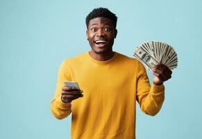Emotional handsome african american young man in yellow using modern cellphone and showing bunch of cash on blue studio background, recommending newest mobile application, copy space photo