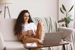 Relaxed pretty young woman watching movie on laptop, drinking coffee, sitting on sofa at home, copy space. Happy curly lady enjoying her weekend alone, watching comedy online, laughing photo