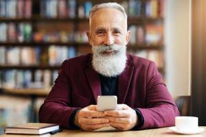 Cheerful elderly grey-haired man with long beard using modern mobile phone while drinking coffee at cafe, chatting with his family or business partners, having notebook by him, free space photo