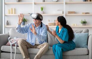 Disabled senior man using VR headset with young nurse, exploring augmented reality at home. Elderly male patient with doctor playing virtual game, having fun with futuristic technologies photo