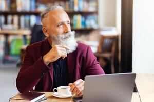 Thoughtful senior man sitting at cafe, using laptop, touching his long beard and looking through window while drinking coffee or tea, copy space. Elderly businessman working online photo