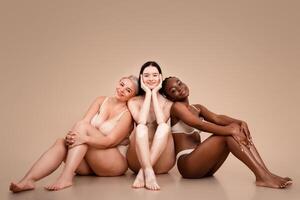 Group of multiethnic women with different body and skin types posing together, attractive multiracial females in underwear sitting on floor over beige background in studio, copy space photo