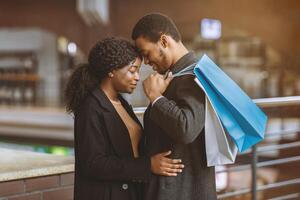 Romance for two, tenderness and date during seasonal sales and Black Friday. Young african american couple in love with colored shopping bags hugging at city mall ready to buy, side view, free space photo