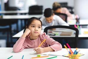 Education And Learning Concept. Portrait of tired and bored small asian girl sitting at desk in classroom at school, writing in notebook and thinking, looking away at window, resting head on hand photo