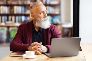 Pensive grey-haired businessman using laptop while having lunch break at cafe, looking through window, copy space. Elderly bearded man working online or websurfing, drinking coffee photo