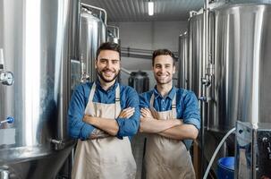 Happy employees at favorite job, small business and male work. Young cheerful men workers in aprons with crossed arms look at camera in interior of factory with large boilers, free space photo