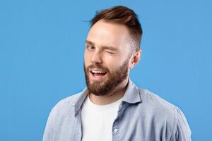 Confident handsome Caucasian man winking and smiling at camera on blue studio background. Playful young bearded guy in casual wear flirting, expressing friendly attitude photo