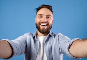 Joyful bearded guy taking selfie on blue studio background. Positive millennial man making photo of himself, smiling at camera, having fun while doing mobile photography