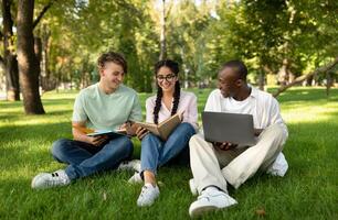 Group of diverse students friends learning, reading books, using laptop and discussing something in university campus park, sitting on grass outside photo