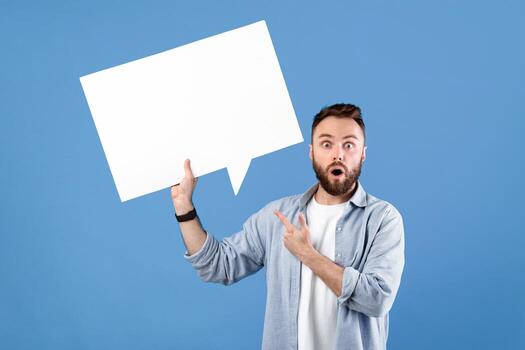 Shocked young guy pointing at empty speech bubble with space for your design over blue studio background, mockup. Attractive bearded man with blank word cloud. Chat, dialog, announcement concept photo