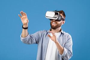 Happy bearded young man using VR headset to explore augmented reality on blue studio background. Cheerful millennial guy playing virtual online game, having fun with modern technologies photo
