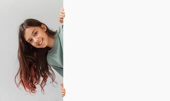 Happy Preteen Girl Posing Hiding Behind Empty White Board And Smiling To Camera In Studio Over Gray Background. Child Holding Paper Poster Advertising Your Text. Panorama, Mockup photo