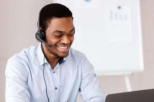 Cheerful black guy crisis manager or customer service representative working in front of laptop in office, wearing headset, having online conversation with clients or business partners, copy space photo