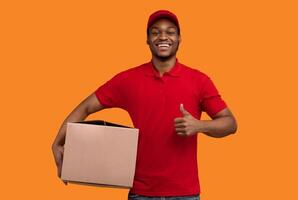 Best Service Concept. Smiling black delivery man wearing red cap and uniform holding cardboard box and showing thumbs up gesture standing isolated over yellow orange studio background photo