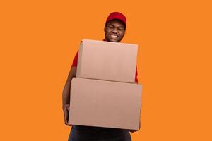 Portrait of african american delivery man wearing red cap and uniform holding stack pile of heavy cardboard boxes, carrying packages with weight stuff, standing isolated over orange studio background photo