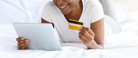 Happy pretty african american woman making order from home, using digital tablet while laying on bed, copy space. Joyful black lady holding credit card, using mobile application for e-commerce on pad photo