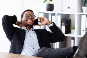 Relaxed black manager sitting with legs on workdesk, having conversation on mobile phone and smiling, talking to his lover while having break at work, resting after successful deal, free space photo