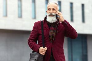 Stylish senior man with long beard talking on mobile phone while comming back home from office, having business conversation, wearing burgundy suit and carrying leather bag, copy space photo