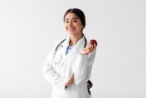 Happy pretty young indian woman doctor dentist in white coat with stethoscope hold fresh red apple on light background. Proper nutrition, vitamins, healthy diet, recommendation for perfect teeth photo