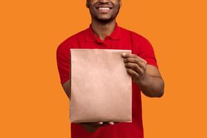 Closeup of unrecognizable smiling african american delivery man in red uniform holding and giving craft paper bag with products from restaurant to camera, isolated on orange studio background photo