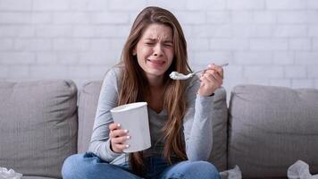 Emotional eating concept. Stressed millennial lady crying on sofa with bucket of ice cream, suffering from relationship problem or watching sad movie, feeling lonely and depressed photo