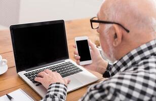 Senior man using laptop and smartphone with blank screen back view, mockup. Mature male surfing net with computer and mobile at home, copy space photo