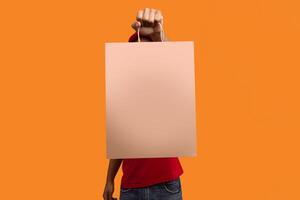 Closeup of african american delivery man in red uniform holding and showing craft paper bag to camera, isolated on orange background. Design, mockup, blank space on package for advert photo