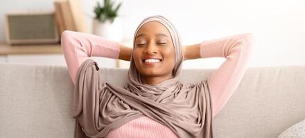 Beautiful Muslim woman in traditional hijab relaxing on sofa with closed eyes, indoors. Smiling black woman in Arab headscarf enjoying her break, taking time off daily routine, resting on couch photo