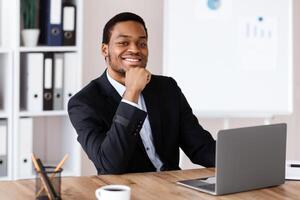 Successful african american businessman sitting in his office, using modern laptop, drinking coffee, smiling at camera, copy space. Cheerful black entrepreneur starting his day with reading emails photo