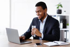 Smiling black manager reading sales report on laptop in his modern office, drinking water. Happy african american manager planning his day, chatting with clients on internet, empty space photo