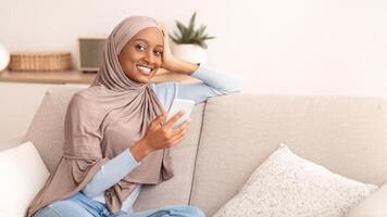 Young black woman in hijab using smartphone for work or studies on sofa at home. Happy Muslim lady browsing web, chatting online or posting in social media on mobile phone photo