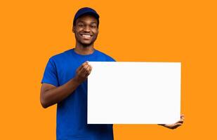 Shipment Service Promotion. Portrait of smiling bearded black delivery man in blue uniform holding and showing square empty blank board with template isolated on orange studio background photo