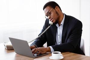 Busy black crisis manager talking on phone with client or colleague and using modern laptop, checking data online, having cup of coffee on his workdesk, office interior, empty space photo