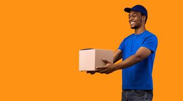 Delivery Service Concept. Smiling black male courier wearing blue cap and uniform holding cardboard box, showing and giving it to the side, copy space, banner. Guy standing on yellow studio background photo