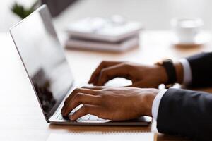 Side view of unrecognizable black manager typing on modern laptop, sending emails or working on business project, office interior, cropped. African american businessman hands on keyboard, closeup photo