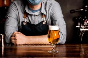Quarantine, nightlife and pub. Bartender in protective mask leans on bar counter on which stands glass of light beer with foam, cropped photo