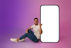 Full length of cheerful young Arab man sitting near big cellphone with mockup for mobile app on screen in neon light. Smartphone display template for new website or online ad photo