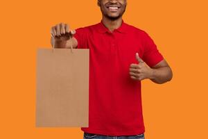 Closeup of unrecognizable black man in red t-shirt holding craft paper bag, showing thumbs up gesture sign, isolated on orange background. Design, mockup, blank space on package for advert photo