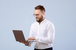 Positive young man in office wear using laptop computer over grey studio background. Joyful millennial guy working or studying remotely, having online business meeting on pc photo