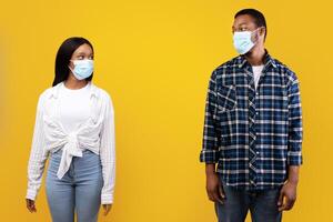 Relationships and social distancing during covid-19 lockdown. Serious young african american students guy and lady in protective masks looking at each other, isolated on yellow background, empty space photo