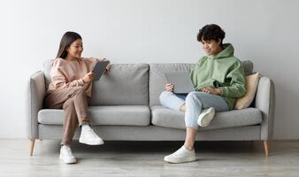 Handsome young Asian guy and his girlfriend using modern gadgets while sitting on couch at home, copy space. Full length of millennial couple communicating online via laptop and tablet pc photo